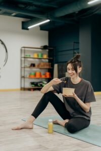 A smiling woman sitting on a mat enjoying a healthy meal indoors after exercise.