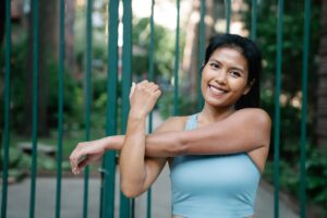 A woman in fitness attire stretching her arms outdoors with a confident smile.