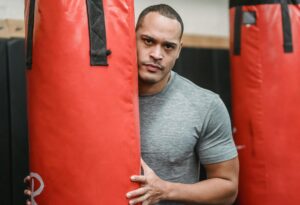 Adult serious ethnic male athlete with muscles in t shirt looking at camera between punching bags in gymnasium