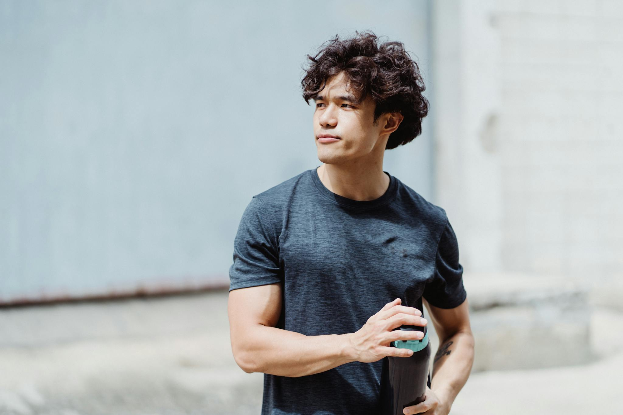 Portrait of a fit young man in a T-shirt holding a fitness bottle outdoors, looking thoughtful.